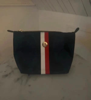 Tommy Hilfiger marinblå necessär  - Snygg marinblå från Tommy Hilfiger med klassiska rödvita ränder framtill och guldfärgad logodetalj.  har dragkedja upptill och stilren form, perfekt för att hålla koll på dina essentials.