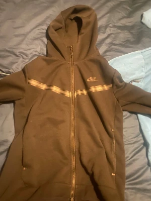 Svart Nike hoodie med dragkedja - Svart hoodie från Nike i storlek M med hel dragkedja och huva. Tröjan har ett diskret mönster med två vågiga linjer över bröstet och Nike-logga på vänster sida. Tillverkad i mjukt bomullsmaterial, perfekt för chill eller träning. Nike tech