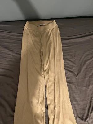 Beige ribbade wide byxor - Säljer ett par beige ribbade byxor med bred resår i midjan. Byxorna har en skön wide fit och är tillverkade i ett mjukt, stretchigt syntetmaterial. Perfekta för dig som gillar avslappnad stil och vill ha något som är både bekvämt och trendigt.