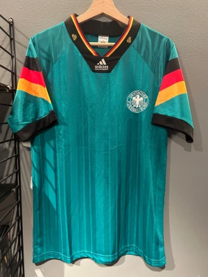 Tysklands retro fotbollströja Adidas - Retro fotbollströja från Adidas med Tysklands landslagsemblem. Tröjan är turkos med subtila vertikala ränder, har svart krage med röd och gul detalj, samt korta ärmar med svarta, röda och gula band. Materialet är glansigt polyester, perfekt för dig som gillar klassisk sportstil. Helt ny med tagg en replika