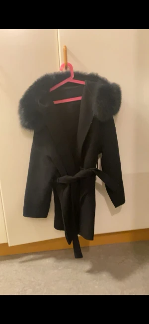 Svart kappa med päls och bälte - Snygg svart kappa med fluffig päls runt kragen och ett matchande bälte i midjan. Jackan har en stilren och rak form, perfekt för dig som gillar att sticka ut med detaljer. Pälsen ger en lyxig vibe och bältet gör att du kan justera passformen.