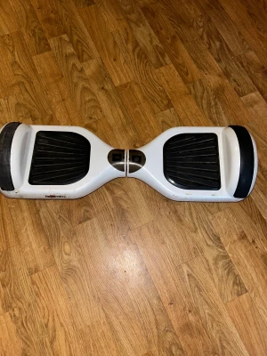 Airboard Hoverboard - Självbalanserande Airboard hoverboard i vitt med svarta fotplattor. Produkten visar tydligt slitage, repor och smuts på undersidan och sidorna, men verkar vara i fungerande skick. Perfekt för dig som vill ha ett prisvärt elfordon för kortare transporter eller nöje. Den fungerar som den ska 