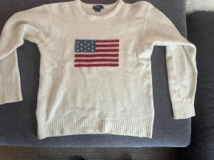 Vit stickad tröja Polo Ralph Lauren - Vit stickad tröja från Polo Ralph Lauren med amerikansk flagga broderad på bröstet och diskret Polo-text. Rund hals, ribbade muddar och långärmad. Perfekt för dig som gillar klassisk stil med en twist.