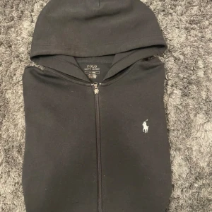 Svart hoodie från Polo Ralph Lauren - Svart hoodie från Polo Ralph Lauren med dragkedja och klassisk vit logga på bröstet. Tröjan är i storlek M och är i bra skick för utom den minimala slitningen vid botten