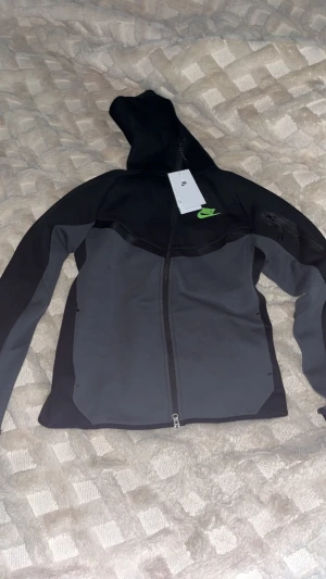 Nike Tech  - Nike vindjacka i svart och mörkgrå med huva och hel dragkedja framtill. Jackan har en sportig look med grönt Nike-logga på bröstet och praktiska fickor. Materialet är mjukt och syntetiskt, perfekt för aktiva dagar. Snygg passform och stilren design.