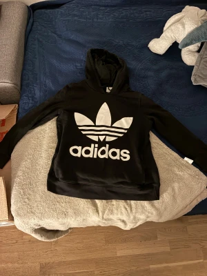 Svart Adidas hoodie med stor logga - Svart hoodie från Adidas med stor vit trefoil-logga och text framtill. Tröjan har huva med snörning och långa ärmar. Materialet är mjuk bomull och polyester, perfekt för chill dagar. Passformen är loose och modellen är klassisk streetwear.