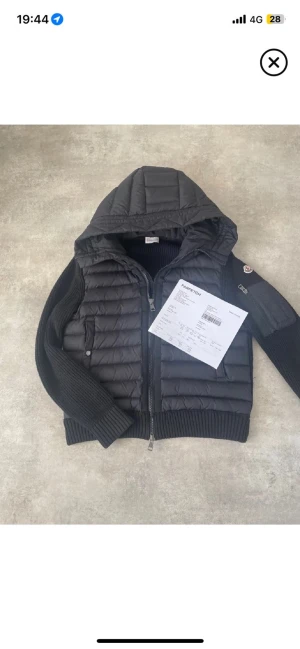 Moncler  kardigan - Moncler kardigan – Storlek S Säljer en exklusiv och eftertraktad Moncler-kardigan i svart. Modellen kombinerar quiltad dunfront med stickade ärmar och sidor – perfekt mix av värme, komfort och premiumstil. Jackan har avtagbar huva, tvåvägsdragkedja och den ikoniska Moncler-patchen på armen. Äkta vara