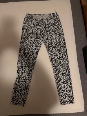 Leopardmönstrade leggings i svart och vitt - Snygga leggings med leopardmönster i svart, vitt och små rosa detaljer. De har en tight passform och är perfekta för dig som vill sticka ut med ett coolt djurmönster. Materialet känns mjukt och stretchigt, vilket gör dem sköna att bära hela dagen.