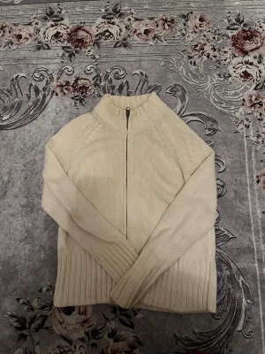 Beige stickad kofta Marks & Spencer - Snygg beige stickad kofta från Marks & Spencer med ribbad krage och muddar. Koftan har hel dragkedja framtill och långärmad design. Perfekt för lager-på-lager och chill dagar. Materialet är mjukt och håller dig varm under kyliga månader.