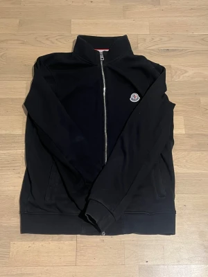 Svart Moncler bomberjacka med dragkedja - Svart bomberjacka från Moncler med klassisk ståkrage och silverfärgad dragkedja framtill. Jackan har ribbade muddar, två sidofickor och Moncler-logga på bröstet. Tillverkad i mjuk bomull för en clean och stilren look.