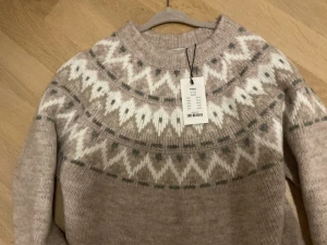 Beige stickad tröja med mönster - Säljer en beige stickad tröja med vitt och grått nordiskt mönster runt halsen. Tröjan har lång ärm, ribbade muddar och rund hals. Perfekt för dig som gillar mysiga och snygga vinterplagg.