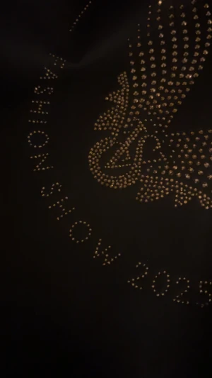 Svart shoppingväska Victoria's Secret - Stor svart shoppingväska från Victoria's Secret med Fashion Show 2025-tryck i guldiga nitar och ikoniska änglavingar på framsidan. Väskan har breda handtag och är gjord i slitstark polyester, perfekt för att bära med sig allt man behöver.