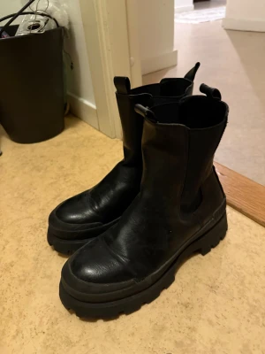 Svarta chunky chelsea boots - Säljer ett par svarta chelsea boots med grov platt sula och rund tå. Bootsens ovandel är i skinn och har elastiska paneler på sidorna för enkel påtagning. Perfekt för dig som gillar chunky stil och vill ha ett par boots som sticker ut.