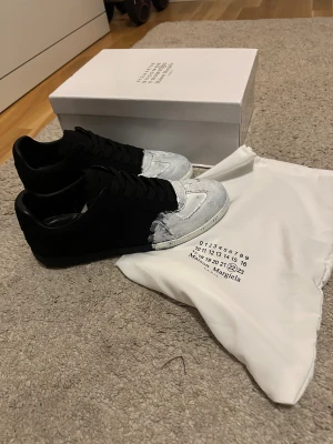 Maison Margiela svartvita sneakers - Maison Margiela sneakers med unik svart och vit färgblockad design. Skorna har snörning, platt sula och rund tå. Materialet är canvas och skinn, med en cool handmålad look på tån och sidorna. Märkets logga syns på sulan och dustbag ingår.KAN GÅ NED I PRIS!!
