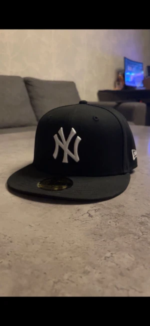 Svart New Era Yankees keps - Snygg svart New Era keps med New York Yankees-logga i vitt framtill och MLB-logga bak. På sidan finns en vit New Era-symbol och en röd 100-emblem. Platt skärm och klassisk form, tillverkad i bomull. Perfekt för dig som vill ha streetstyle med sportig vibe.