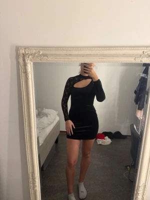 Svart sammetsklänning med spetsärm - Supersnygg svart sammetsklänning med en unik cut-out vid halsen och en lång spetsärm. Den är tight och kort, vilket ger en riktigt trendig look. Perfekt för dig som vill sticka ut med en cool detalj och älskar kombon av sammet och spets.