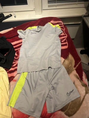 Grå träningsshorts från Nike - Snygga grå träningsshorts från Nike med en fräsch neongul sidopanel och klassisk Nike-logga på benet. Shortsen har snörning i midjan och är gjorda i ett lätt och ventilerande polyestermaterial, perfekta för sport eller gym.