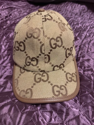 Gucci beige keps med GG-mönster - Snygg beige keps från Gucci med det klassiska GG-mönstret i brunt över hela kepsen. Kepsen har bruna detaljer och justerbar rem baktill i skinn med Gucci-logga. Materialet är en mix av bomull och syntet, perfekt för dig som vill ha en lyxig streetstyle-look.