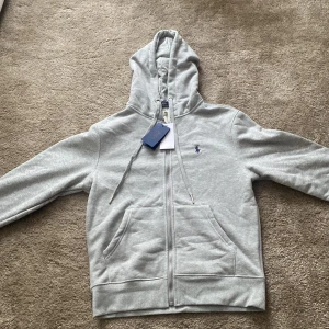 Polo Ralph Lauren hoodie - Snygg grå hoodie från Polo Ralph Lauren med dragkedja och huva. Sprillans ny säljer pga att den var liten i storleken hör av er vi minsta fråga!  PS- jag är 184 så om du är längre än det så skulle jag inte rekommendera denna. (It äkt)