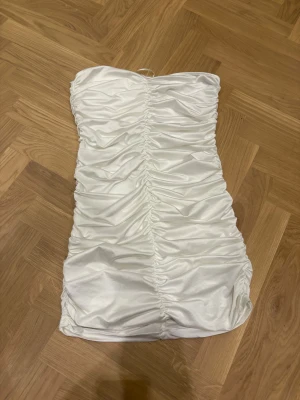 Vit omlott kortklänning från Divided - Supersnygg vit kortklänning från Divided i storlek L. Klänningen är axelbandslös och har ett glansigt satinliknande material med snygg rynkning framtill som ger en cool och trendig look. Perfekt för dig som vill sticka ut med en chic och modern stil.