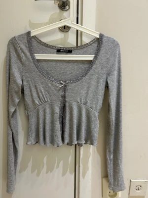 Grå croppad topp med spets från Gina Tricot - Supersöt ljusgrå croppad topp från Gina Tricot med lång ärm. Rundad halsringning med spetskant och liten rosett framtill. Toppens material är mjuk bomull och den har en lätt rynkad effekt under bysten för en feminin siluett.