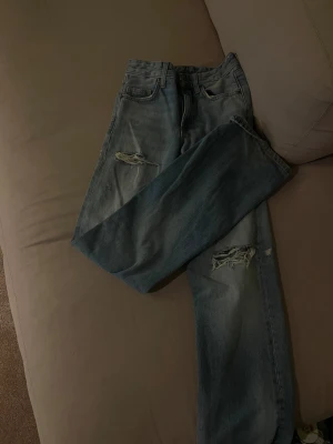 Blå ripped jeans från Zara - Snygga blå jeans från Zara med slitna detaljer och hål vid knäna. Modellen har fem fickor, bälteshällor och klassisk knappstängning. Jeansen har en rak passform och är tillverkade i bomull för en skön känsla. Dom är användbara 1 gång och inga deferenser.