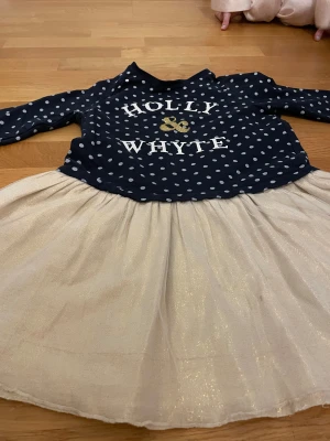 Prickig klänning Holly & Whyte 128 - Supersöt klänning från Holly & Whyte by Lindex i storlek 128. Mörkblå överdel med vita prickar och text, samt glittrig ljusbeige kjol. Långärmad och perfekt för dig som gillar lekfull stil med lite extra glitter.