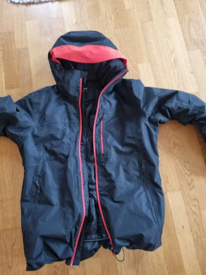 Svart Everest vindjacka med röd detalj - Svart vindjacka från Everest med röd insida på huvan och röda detaljer längs dragkedjan. Jackan har justerbara muddar med kardborre, flera fickor med dragkedja och en stor huva. Perfekt för blåsiga dagar och outdoor-aktiviteter.