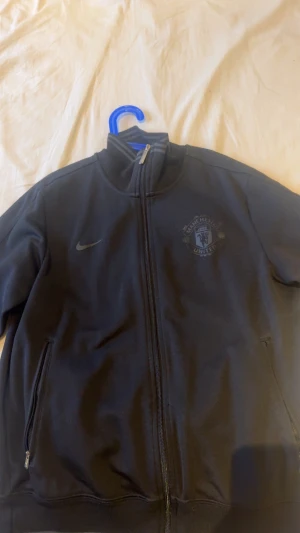 Manchester United svart vindjacka Nike - Svart tjocktröja från Nike med Manchester United-logga på bröstet och stor text på ryggen. Tröjan har hel dragkedja, ståkrage och två fickor med dragkedja. Perfekt för dig som vill representera ditt lag med stil. Använd 2 gånger.