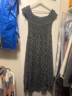 Svart blommig maxiklänning från MIS - Svart långklänning från MIS med vita småblommor, smockad överdel och korta ärmar. Klänningen har en söt, lätt utställd kjol och elastisk passform upptill. Perfekt för dig som gillar romantiska och bekväma plagg med sommarkänsla.