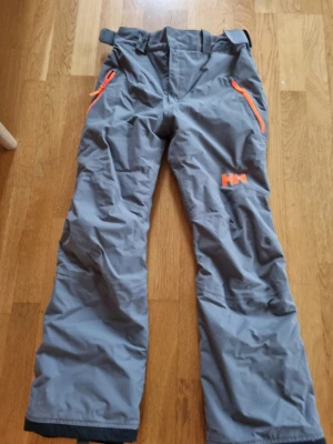 Grå täckbyxor från Helly Hansen - Säljer ett par grå täckbyxor från Helly Hansen med orange detaljer och dragkedjor. Byxorna har justerbar midja, förstärkta benslut med snölås och är tillverkade i slitstarkt syntetmaterial. Perfekta för kalla dagar och utomhusaktiviteter.