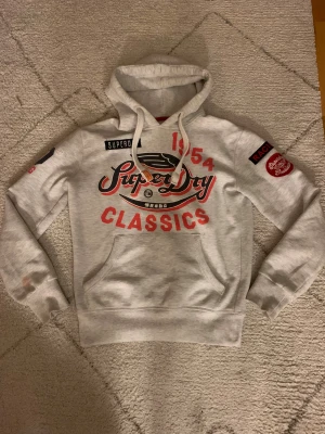 Vit Superdry hoodie med tryck - Snygg vit hoodie från Superdry med stort rött och svart tryck på bröstet och coola broderade detaljer på ärmen. Tröjan har huva med snörning och en klassisk känguruficka framtill. Perfekt för dig som gillar streetwear och sportig stil.