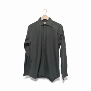 Svart skjorta från John Henric - Stilren svart skjorta från John Henric med klassisk krage och knappar framtill. Skjortan är långärmad och har en snygg, rak passform. Perfekt för dig som gillar enkel och clean stil. Materialet är mjukt och känns skönt mot huden.