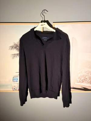 Svart långärmad polotröja Riley - Svart polotröja från Riley med klassisk krage och v-ringad öppning. Tröjan är långärmad och har ribbade muddar vid ärmslut och nederkant. Tillverkad i mjukt bomullsmaterial som känns skönt mot huden. Perfekt för dig som gillar stilrena och enkla plagg. 