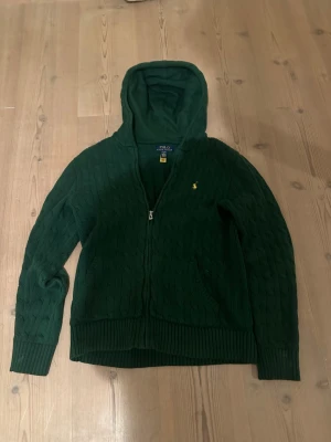 Grön stickad hoodie från Polo Ralph Lauren - Mörkgrön stickad hoodie från Polo Ralph Lauren med dragkedja och huva. Tröjan har kabelstickat mönster, ribbade muddar och fickor framtill. Ikoniska Polo-loggan i gult på bröstet. Perfekt för chill dagar och snygg till jeans.