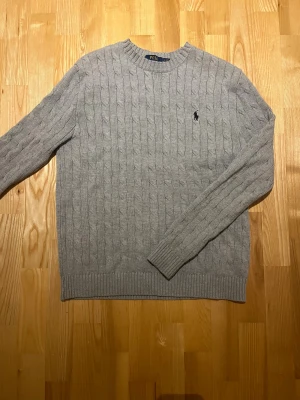 Grå stickad tröja från Polo Ralph Lauren - Snygg grå kabelstickad tröja från Polo Ralph Lauren med klassisk rund hals och broderad logga på bröstet. Tröjan har ribbade muddar vid ärmslut och nederkant, och är långärmad. Perfekt för dig som gillar stilrena och tidlösa plagg.