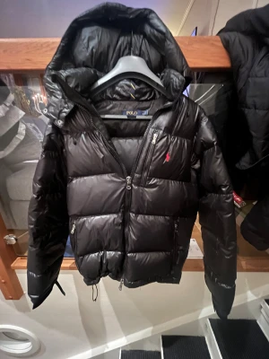 Svart pufferjacka från Polo Ralph Lauren - Svart pufferjacka från Polo Ralph Lauren med huva och dragkedja framtill. Jackan har två sidofickor med dragkedja och den klassiska röda Polo-loggan på bröstet. Materialet är glansigt och fylligt, perfekt för kalla dagar. Pris går att diskuteras.