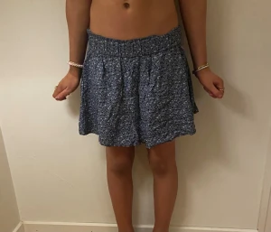 Blå blommig kortkjol med volang - Supersöt blå kortkjol med småblommigt mönster och volangkant. Kjolen har en bred resår i midjan som gör den bekväm och lätt att bära. Perfekt för dig som gillar en avslappnad och feminin stil.