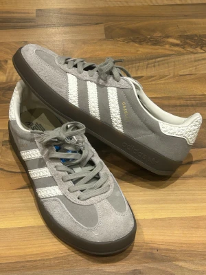 Grå Adidas Gazelle sneakers mocka - Ett par grå Adidas Gazelle sneakers med klassiska vita ränder och detaljer i guld. Ovandelen är i mjuk mocka och har snörning. Sulan är brun med mönstrad undersida och Adidas-loggor. Skorna har en rund tå och platt sula för skön komfort.