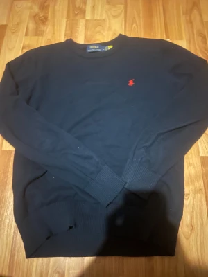 Mörkblå tröja från Polo Ralph Lauren - Klassisk mörkblå tröja från Polo Ralph Lauren med rund hals och röd broderad logga på bröstet. Tröjan har ribbade muddar vid ärmslut och nederkant. Perfekt för dig som gillar stilren och tidlös design.
