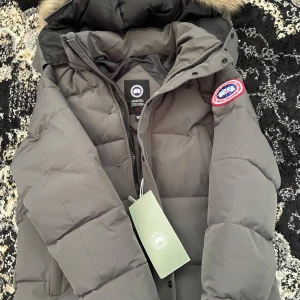  Canada Goose  - Säljer en grå dunjacka från Canada Goose med klassisk patch på ärmen och pälskantad huva. Jackan är quiltad, har dragkedja och tryckknappar framtill. Perfekt för kalla vinterdagar och har en clean, stilren look.