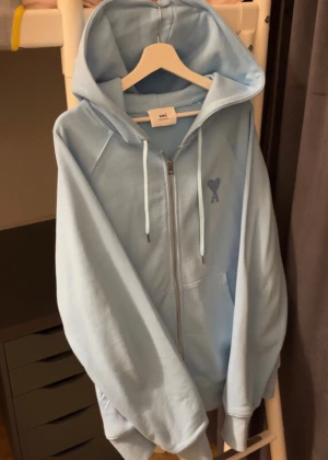 Ljusblå zip hoodie från Ami Paris - Säljer en ljusblå hoodie från Ami Paris med dragkedja och huva. Tröjan har broderad logga med hjärta och A på bröstet, snörning i huvan och känguruficka. Tillverkad i mjuk bomull, perfekt för chill dagar.