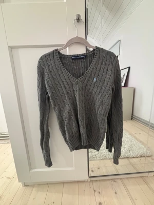Grå kabelstickad tröja Ralph Lauren - Grå kabelstickad tröja från Ralph Lauren Sport med v-ringad hals och lång ärm. Klassisk design med broderad logga i ljusblått på bröstet. Ribbad kant vid hals, ärmslut och nederkant. Perfekt för dig som gillar stilrena och tidlösa plagg.