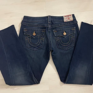 True Religion mörkblå bootcut jeans - Snygga mörkblå jeans från True Religion med klassiska kontrastsömmar och ikoniska fickdetaljer bak. Modellen heter joey och har bootcut-ben och låg midja, tillverkade i slitstarkt denim med guldfärgade knappar och nitar. Perfekt för dig som gillar statement-jeans. Det är bara att höra av dig vid frågor!💕 Midjemått på sida 5 och innerbenslängden på sida 6