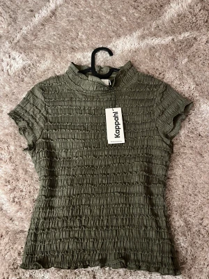 Grön ribbad topp från Kappahl - Säljer en grön ribbad topp från Kappahl med korta ärmar och hög krage. Toppen har ett smockat, stretchigt tyg som ger en cool textur och sitter snyggt på kroppen. Perfekt för dig som gillar trendiga detaljer och enkel stil. Aldrig använd