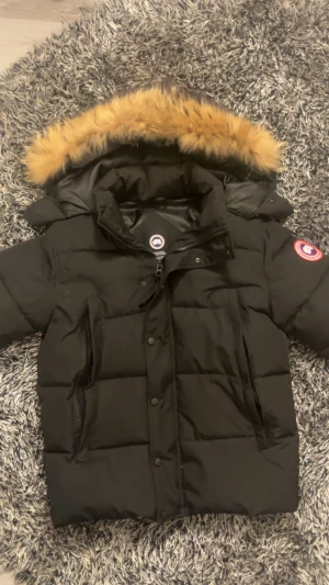 Svart dunjacka från Canada Goose - Säljer en svart dunjacka från Canada Goose med ikonisk patch på ärmen och stor huva med beige fuskpäls. Jackan har dragkedja, knappar och flera praktiska fickor. Perfekt för kalla vinterdagar och riktigt snygg streetstil. Det passar också S