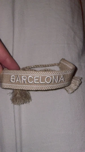 Beige armband med Barcelona-tryck - Snyggt beige armband med texten 'BARCELONA' broderad i vitt. Armbandet har fransar i ändarna och är gjort i ett grovt tygmaterial med flätad knytning. Perfekt accessoar för dig som gillar unika detaljer och vill visa din kärlek till Barcelona.