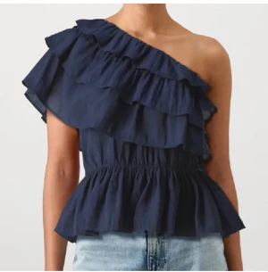 Marinblå volang topp oneshoulder - Säljer en marinblå topp med volanger och oneshoulder-design. Toppen har en snygg, asymmetrisk axel och är tillverkad i ett lätt material som ger en luftig känsla. Perfekt för dig som gillar att sticka ut med coola detaljer och volanger.