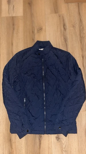 Mörkblå quiltad pufferjacka - Snygg mörkblå pufferjacka med quiltad design och dragkedja framtill. Jackan har ståkrage, två sidofickor med dragkedja och knappdetaljer vid ärmslut. Perfekt för dig som gillar stilren och sportig look.