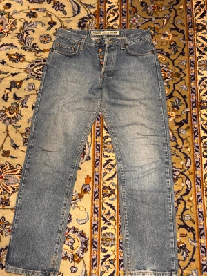 Blå jeans från Solo Denim, storlek 32/32 - Säljer ett par klassiska blå jeans från Solo Denim, modell 754. Jeansen har raka ben, fem fickor och knäppning med fyra metallknappar framtill. Tillverkade i 100% bomull för en autentisk denimkänsla. Perfekt för dig som gillar tidlös stil och schysst passform.